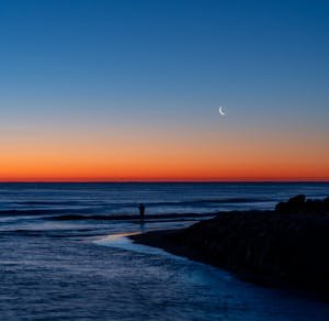 A tranquil twilight scene of a beach in Valencia, Spain, with a crescent moon.